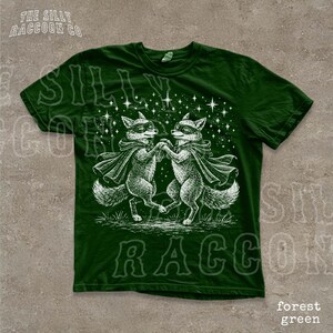 A green t-shirt with a white illustration of two foxes dancing together, surrounded by stars.