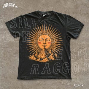 A black t-shirt with a graphic design depicting a sun with rays emanating from it, surrounded by mountains.
