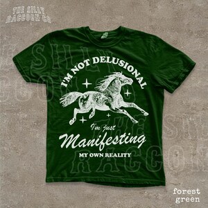 A green t-shirt with a graphic design depicting a horse in mid-gallop and the text "I'm not delusional, I'm just Manifesting, MY OWN REALITY" printed on it.