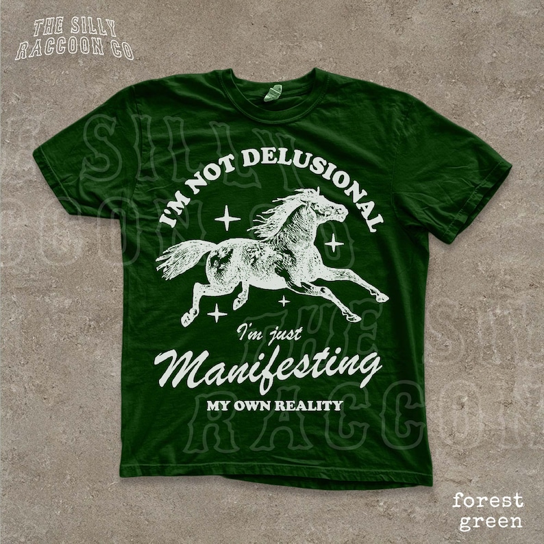 A green t-shirt with a graphic design depicting a horse in mid-gallop and the text "I'm not delusional, I'm just Manifesting, MY OWN REALITY" printed on it.
