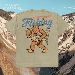 a t-shirt with a cartoon depiction of a giant, furry creature holding a fish and wearing a fishing hat. The t-shirt has a humorous message printed on it, which says "I'D Rather Be Fishing".