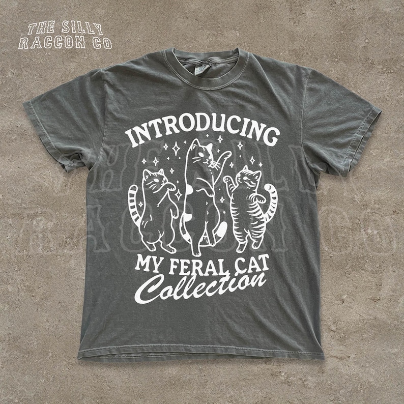 A gray t-shirt with a graphic design featuring three cats and the text "INTRODUCING MY FERAL CAT COLLECTION" printed on it.