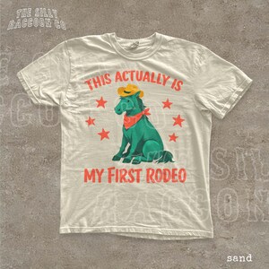 A white t-shirt with a graphic design featuring a green cartoon horse wearing a cowboy hat and a red bandana, surrounded by red stars. The text on the t-shirt reads "THIS ACTUALLY IS MY FIRST RODEO".