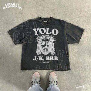 A black t-shirt with a graphic design and text printed on it, along with a pair of sneakers and a person's feet.