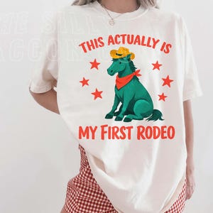 A woman wearing a white t-shirt with a graphic of a green horse and the text "This actually is my first rodeo" printed on it.