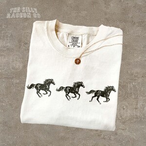 A white t-shirt with a graphic design of three black horses running in a line, along with a small pendant necklace.