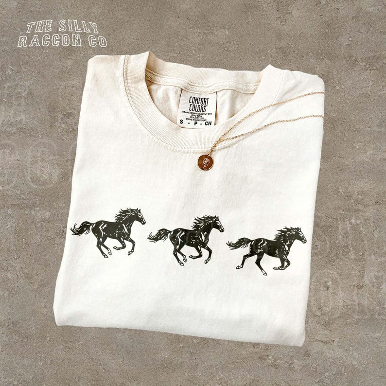 A white t-shirt with a graphic design of three black horses running in a line, along with a small pendant necklace.