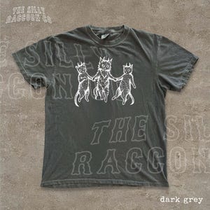 A gray t-shirt with a graphic design featuring three cats with horns and a cat with a lion's head, along with the text "The silly raccoon co" printed on it.