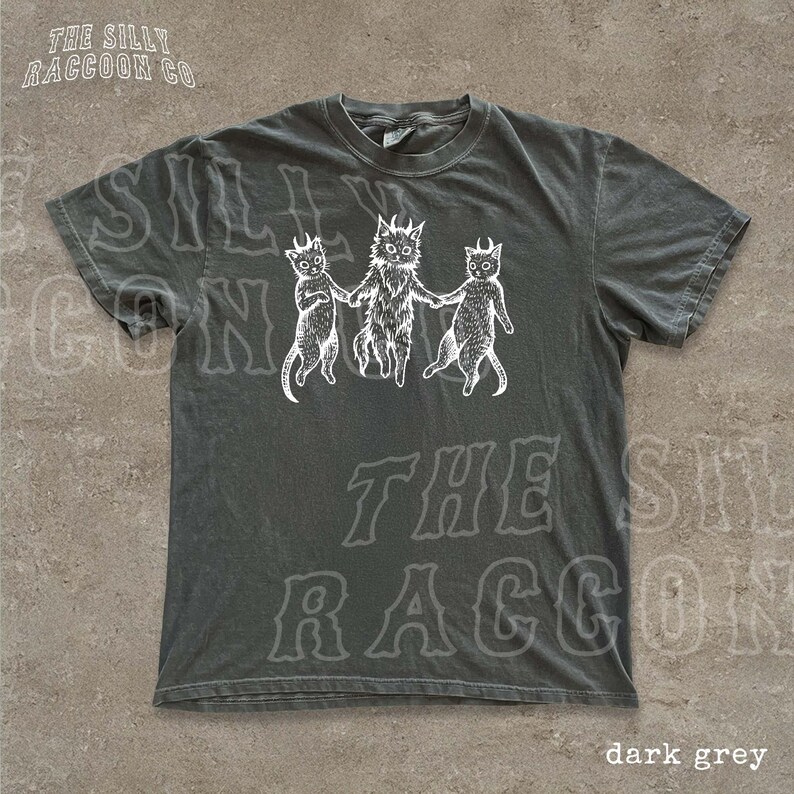 A gray t-shirt with a graphic design featuring three cats with horns and a cat with a lion's head, along with the text "The silly raccoon co" printed on it.