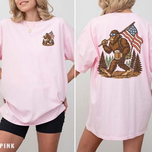 A woman wearing a pink t-shirt with an illustration of a Sasquatch carrying an American flag.