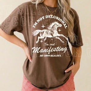 A woman wearing a brown t-shirt with a graphic design and text, standing against a plain background.