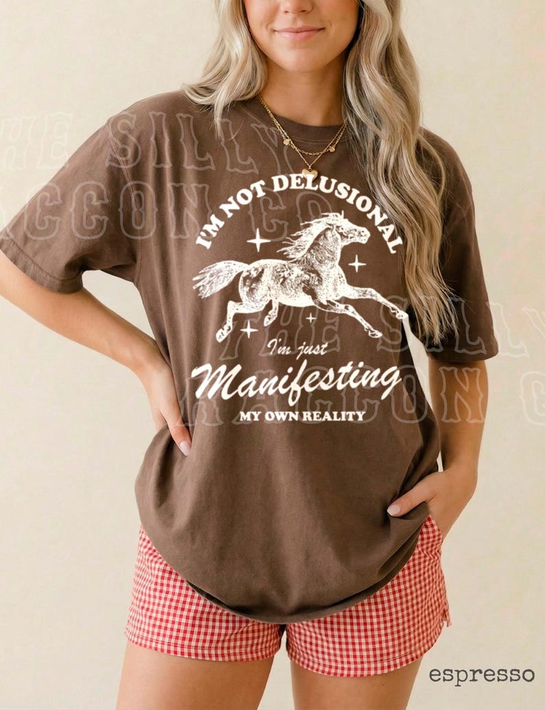 A woman wearing a brown t-shirt with a graphic design and text, standing against a plain background.