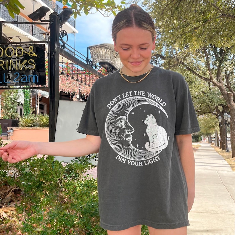 Celestial moon t-shirt featuring a vintage-style crescent moon with a human face and a cosmic black cat sitting on top. Astrology-inspired design with a witchy aesthetic, mystical starry details, and a motivational quote. Soft cotton blend, unisex fit, and available in various sizes. Perfect for astrology lovers, witchcore fashion fans, and those who adore celestial art. Great gift for spiritual souls, moon phase enthusiasts, and black cat lovers.