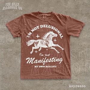 a brown t-shirt with a graphic design of a horse and the text "I'm not delusional, I'm just Manifesting my own reality" printed on it.
