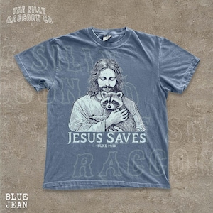 A t-shirt design featuring an illustration of Jesus holding a raccoon, with the text "JESUS SAVES" and "LUKELINE 1910" printed on it.