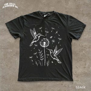 A black t-shirt with a white graphic design depicting two hummingbirds hovering over a dandelion surrounded by flowers and plants.