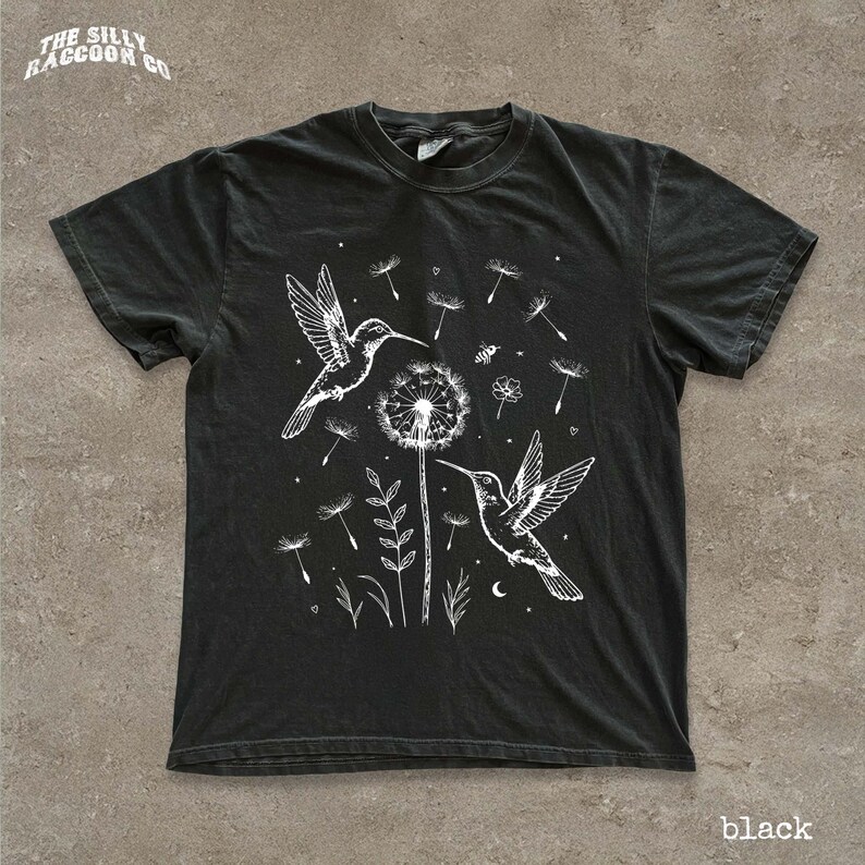 A black t-shirt with a white graphic design depicting two hummingbirds hovering over a dandelion surrounded by flowers and plants.