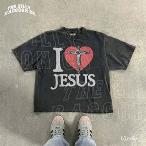 A black t-shirt with a graphic design of a cross and a heart, along with the text "I I love JESUS" printed on it. The t-shirt is laid out on a concrete floor, and a pair of white sneakers is visible underneath it.