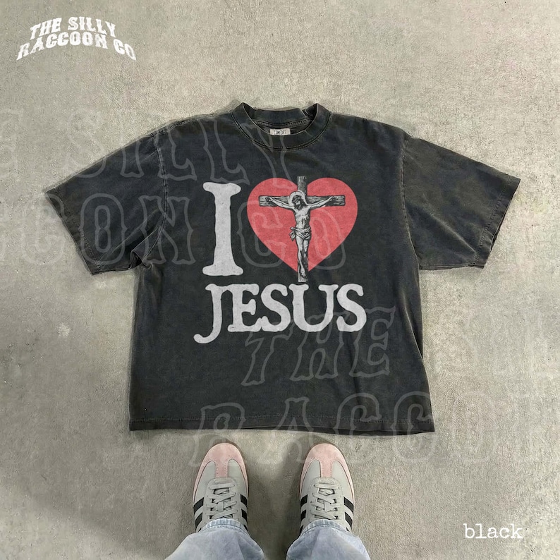 A black t-shirt with a graphic design of a cross and a heart, along with the text "I I love JESUS" printed on it. The t-shirt is laid out on a concrete floor, and a pair of white sneakers is visible underneath it.