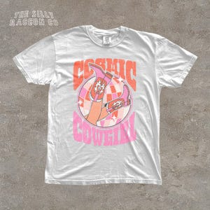 A white t-shirt with a graphic design featuring a pair of cowboy boots and the text "COSMIC COWGIRL" in a retro style.