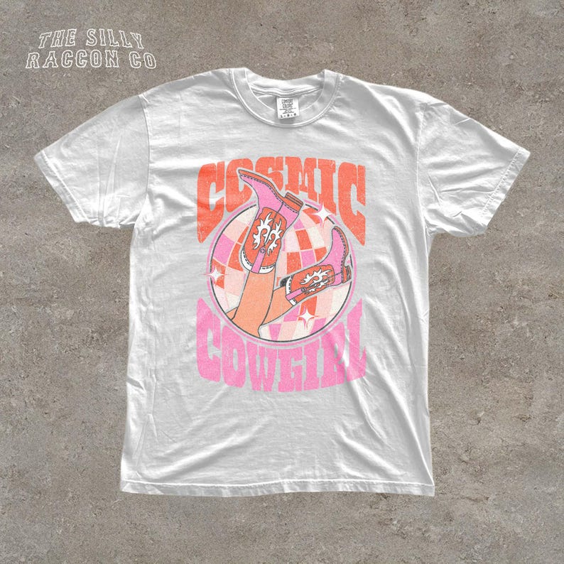 A white t-shirt with a graphic design featuring a pair of cowboy boots and the text "COSMIC COWGIRL" in a retro style.