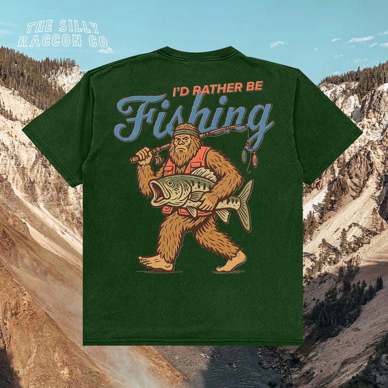 A green t-shirt with a graphic design depicting a large, furry creature holding a fish and the text "I'D Rather Be Fishing" printed on it.