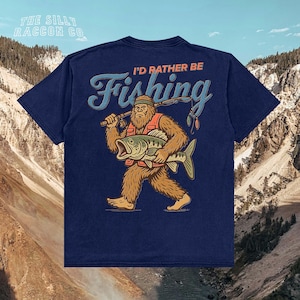 A blue t-shirt with a graphic design depicting a large, furry creature holding a fish and the text "I'D Rather Be Fishing" printed on it.