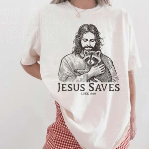 A woman wearing a white t-shirt with a graphic of Jesus holding a raccoon, along with the text "JESUS SAVES" and " Luke 19:10".