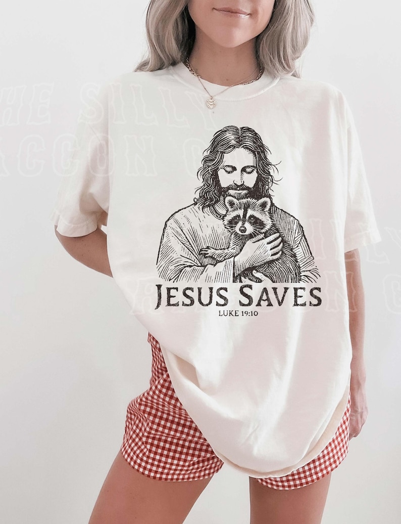 A woman wearing a white t-shirt with a graphic of Jesus holding a raccoon, along with the text "JESUS SAVES" and " Luke 19:10".