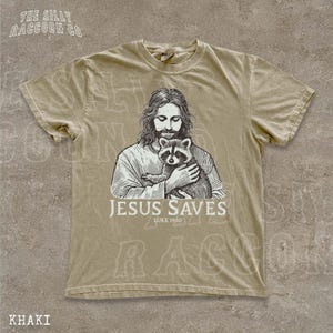 A t-shirt with a graphic design depicting a man holding a raccoon, along with the text "JESUS SAVES LUKELINE 1910" printed on it.