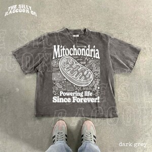 A gray t-shirt with a graphic design depicting Mitochondria, a cellular organelle, and the text "Powering life since forever!" printed on it. The t-shirt is laid out on a concrete floor, and a pair of feet is visible in the bottom right corner of the image.
