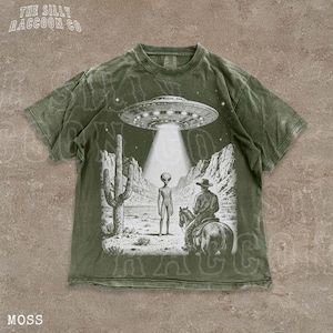 A t-shirt with a graphic design depicting an alien spacecraft hovering over a desert landscape, with a cowboy on a horse below.