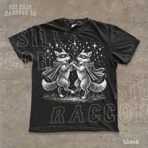 A black t-shirt with a graphic design featuring two foxes dancing under a starry sky.