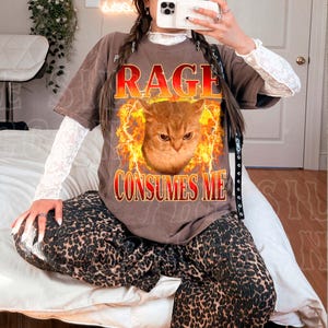 A person is sitting on a bed, wearing a t-shirt with a graphic of a cat and the text "RAGE CONSUMES ME" printed on it.