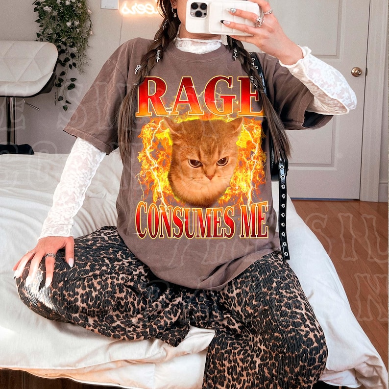 A person is sitting on a bed, wearing a t-shirt with a graphic of a cat and the text "RAGE CONSUMES ME" printed on it.