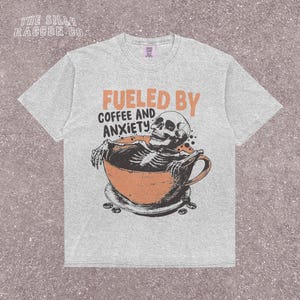Funny coffee shirt for anxious introverts, featuring a skeleton relaxing in a giant coffee cup with the text “Fueled by Coffee and Anxiety.” Vintage grunge design with distressed print and muted tones. Ideal for women, men, and non-binary adults who love dark humor and caffeine culture. Great for Halloween, work-from-home vibes, mental health awareness, or gifting your favorite goth coffee lover.