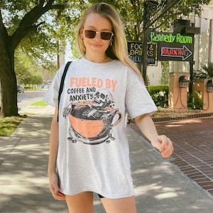 Funny coffee shirt for anxious introverts, featuring a skeleton relaxing in a giant coffee cup with the text “Fueled by Coffee and Anxiety.” Vintage grunge design with distressed print and muted tones. Ideal for women, men, and non-binary adults who love dark humor and caffeine culture. Great for Halloween, work-from-home vibes, mental health awareness, or gifting your favorite goth coffee lover.