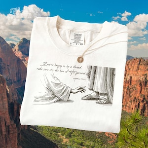 May include: Off-white t-shirt featuring a graphic illustration and text. The illustration shows a hand reaching towards a figure's feet. The text reads, "If you're hanging on by a thread, make sure it's the hem of His garment." A gold necklace is draped on the shirt.