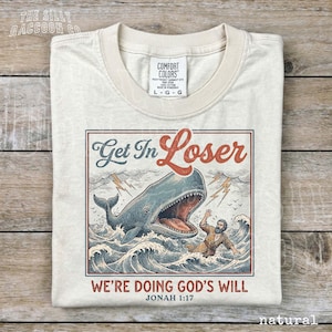 a white t-shirt with a graphic design depicting a man being attacked by a large whale, with the whale's mouth open and the man being held underwater. The text on the t-shirt reads "Get In Loser" and "We're Doing God's Will" along with the Bible verse " Jonah 1:17".
