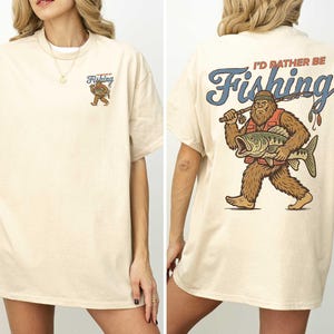 a woman wearing a white t-shirt with a graphic design on the back. The design depicts a large fish with a bear-like appearance, and the text on the back reads "I'd rather be Fishing".