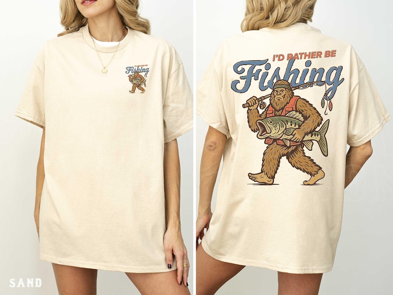 a woman wearing a white t-shirt with a graphic design on the back. The design depicts a large fish with a bear-like appearance, and the text on the back reads "I'd rather be Fishing".