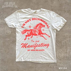A white t-shirt with a graphic design of a red horse and the text "I'm not delusional, I'm just Manifesting my own reality" printed on it.