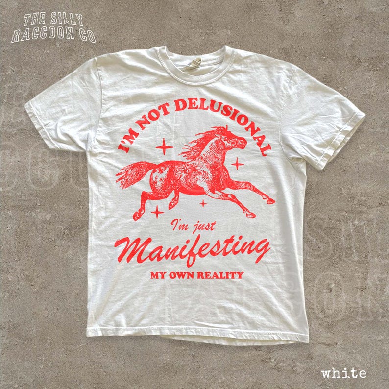 A white t-shirt with a graphic design of a red horse and the text "I'm not delusional, I'm just Manifesting my own reality" printed on it.