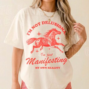 A woman wearing a white t-shirt with a graphic design of a horse and the text "I'm not delusional, I'm just Manifesting my own reality" printed on it.