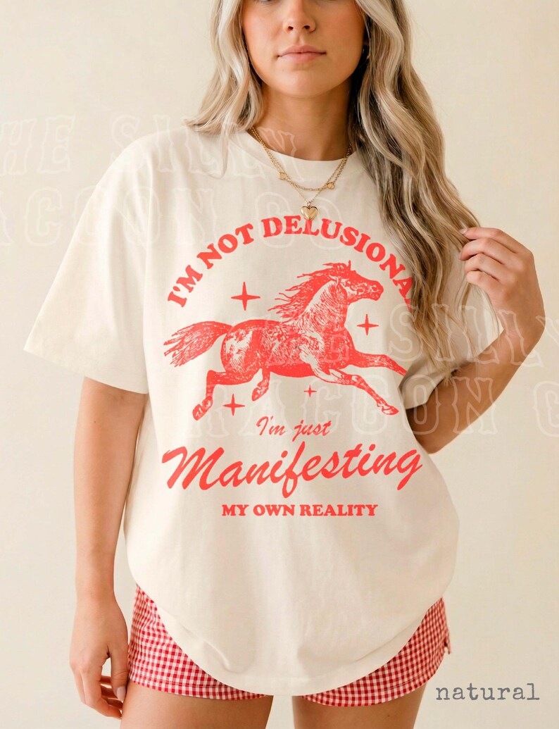 A woman wearing a white t-shirt with a graphic design of a horse and the text "I'm not delusional, I'm just Manifesting my own reality" printed on it.