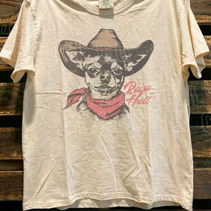 A t-shirt with a graphic design of a dog wearing a cowboy hat and a red scarf, along with the text "Raise Hell" printed on it.