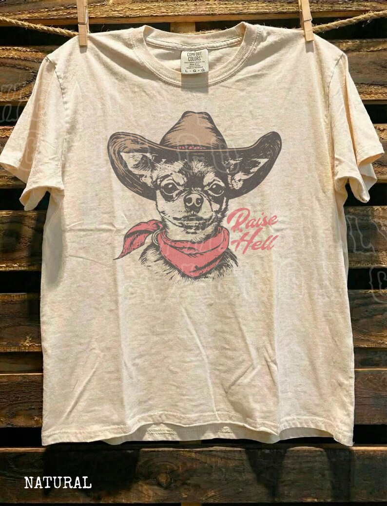 A t-shirt with a graphic design of a dog wearing a cowboy hat and a red scarf, along with the text "Raise Hell" printed on it.