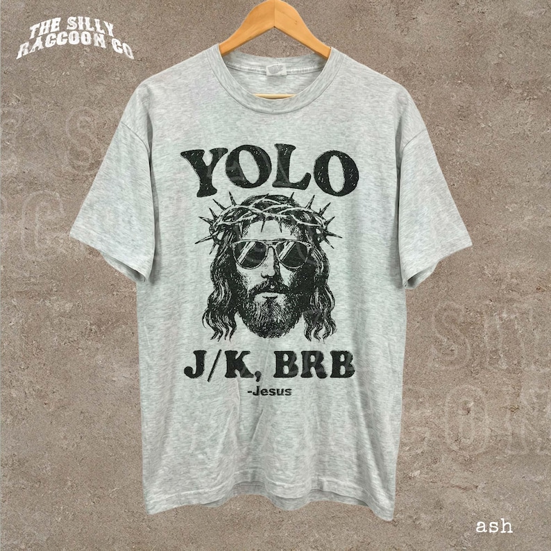 A gray t-shirt with a graphic design featuring a stylized depiction of Jesus wearing sunglasses and a crown of thorns, along with the text "YOLO J/K, BRB - Jesus ash".
