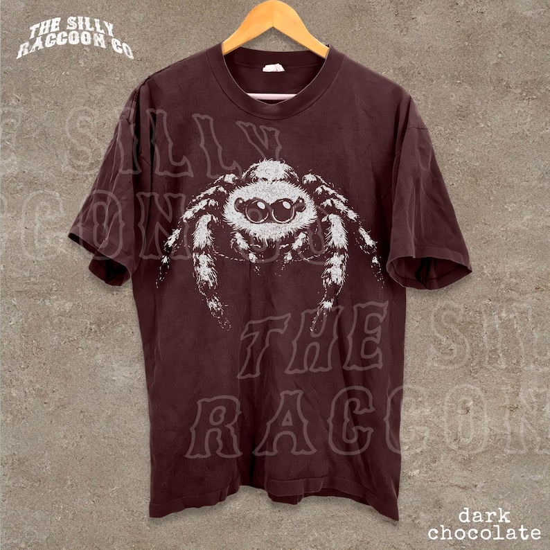 A dark brown t-shirt with a white spider graphic printed on it, hanging on a wooden hanger against a concrete wall.