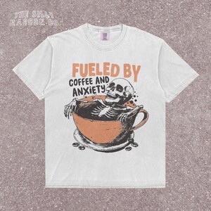 Funny coffee shirt for anxious introverts, featuring a skeleton relaxing in a giant coffee cup with the text “Fueled by Coffee and Anxiety.” Vintage grunge design with distressed print and muted tones. Ideal for women, men, and non-binary adults who love dark humor and caffeine culture. Great for Halloween, work-from-home vibes, mental health awareness, or gifting your favorite goth coffee lover.